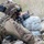 TACP Lightning Challenge 2022 A heavily equipped soldier in multicam camouflage is in a prone firing position behind sandbags, aiming a rifle downrange. The soldier is wearing advanced tactical gear including a helmet with communications equipment, tactical vest, and hearing protection. The setting appears to be an outdoor training range or field exercise.