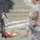 TACP Lightning Challenge 2022 Two military personnel in multicam/OCP camouflage uniforms are conducting what appears to be a field training exercise in an open grassy terrain. The foreground soldier is handling an orange rescue/litter stretcher on the ground, suggesting a casualty evacuation or MEDEVAC training scenario. A second soldier stands in the blurred foreground observing or participating in the exercise.