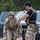 TACP Lightning Challenge 2022 Two military personnel are engaged in what appears to be a training exercise. The soldier in the foreground, wearing a South Korean flag patch and full tactical gear, is carrying two tan military-style water/fuel canisters. A second individual wearing a black shirt labeled 'INSTRUCTOR' is observing and appears to be timing or evaluating the exercise.