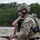 TACP Lightning Challenge 2022 A military personnel wearing full tactical gear, identified by 'TACP' markings on their helmet, is handling a pistol at what appears to be a shooting range or training exercise. The individual is wearing MultiCam camouflage, a ballistic helmet with communications equipment, and carries a medical pouch. A second pistol is visible on the wooden table in front of them.