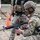 TACP Lightning Challenge 2022 A military personnel wearing TACP (Tactical Air Control Party) designation on their helmet is kneeling and performing a weapons check or reload on an M4/AR-15 style carbine rifle. A second individual in camouflage and tactical vest stands in the background near an orange traffic cone, suggesting a training range environment. The setting appears to be an outdoor military training exercise or qualification course.