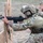TACP Lightning Challenge 2022 A military personnel identified as TACP (Tactical Air Control Party) is in a shooting position behind a wooden barricade, aiming an M4/M16-style rifle. The individual is wearing full multicam combat gear including a tactical helmet with 'TACP' markings, plate carrier vest, and hearing protection. This appears to be a training exercise at an outdoor shooting range.