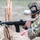 TACP Lightning Challenge 2022 A U.S. Air Force TACP (Tactical Air Control Party) operator is seen in a shooting position, aiming an M4-style carbine rifle around a wooden barrier. The operator is wearing full tactical gear including a multicam helmet labeled 'TACP', communications headset, and body armor. The setting appears to be an outdoor training range or exercise environment.