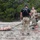 TACP Lightning Challenge 2022 A military or tactical training exercise is underway in an outdoor rocky terrain environment. An instructor wearing a black 'INSTRUCTOR' shirt and hearing protection oversees a trainee dragging a medical training mannequin strapped to a litter/stretcher using a rope or strap. A second observer in camouflage uniform watches and appears to be taking notes or scoring the exercise.