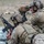 TACP Lightning Challenge 2022 Two military personnel in full combat gear are positioned in a prone/kneeling firing position in an outdoor rocky terrain environment. The forward soldier is aiming a rifle while resting on sandbags, and the rear soldier is using a Leupold spotting scope, suggesting a sniper/spotter team configuration. Both wear MultiCam (OCP) pattern uniforms with tactical helmets, plate carriers, and communications equipment.