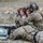 TACP Lightning Challenge 2022 Two military personnel in multicam camouflage uniforms are conducting a shooting exercise in an arid outdoor environment. One soldier is in a prone position operating a rifle supported by sandbags, while a second soldier in a kneeling position uses a Leupold spotting scope or rangefinder. Both are equipped with tactical gear including helmets, body armor, and communication devices.