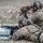 TACP Lightning Challenge 2022 Two military personnel in full combat gear are conducting a shooting exercise at a range. One soldier is in the prone position aiming an M4-style carbine rested on sandbags, while a second soldier in a seated position observes through a Leupold spotting scope mounted on a tripod. Both wear MultiCam (OCP) camouflage uniforms with tactical vests, helmets, and communication headsets.
