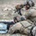 TACP Lightning Challenge 2022 Two military personnel in full combat gear are engaged in a shooting exercise, with one soldier in the prone position firing a rifle supported by sandbags, and a second soldier acting as a spotter using an optic device on a tripod. Both wear MultiCam (OCP) camouflage uniforms with tactical vests, helmets, and communication headsets. The setting appears to be an outdoor range or training area with rocky, arid terrain.