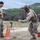 TACP Lightning Challenge 2022 Two military personnel in MultiCam/OCP camouflage uniforms are engaged in what appears to be a training exercise on an open field. One soldier is fully equipped with helmet, body armor, and tactical gear, while the other is in a more relaxed uniform configuration. An orange traffic cone, a camera tripod, and orange equipment markers are visible on the ground.