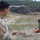 TACP Lightning Challenge 2022 Two military personnel in camouflage uniforms are engaged in what appears to be a field training exercise on an open grassy terrain. One soldier in the foreground is out of focus, while a second soldier in the background is actively dragging or pulling an orange rescue/casualty drag bag across the ground. The setting appears to be a military training range with sparse vegetation and treeline in the background.