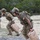 TACP Lightning Challenge 2022 Two military personnel in full tactical gear are carrying water jerry cans while running across a shingle platform on an outdoor training course. Both individuals wear camouflage uniforms, tactical vests, and helmets with communications equipment. The scene appears to be a physical fitness or combat readiness training exercise.