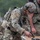 TACP Lightning Challenge 2022 A heavily equipped soldier in MultiCam camouflage uniform is handling a tan/FDE-colored semi-automatic pistol over a field table, appearing to inspect or load the weapon. The soldier is wearing a ballistic helmet with NVG mount, tactical hearing protection, plate carrier with full kit, backpack, and tactical gloves. Rain is visible in the image, suggesting an outdoor training or operational environment.