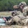 TACP Lightning Challenge 2022 Two military personnel in MultiCam/OCP camouflage uniforms are engaged in a shooting range exercise. One soldier is in the prone position aiming an M4-style carbine resting on sandbags, while a second soldier kneels behind using a Leupold spotting scope. The setting appears to be an outdoor range with green treeline in the background and light rain visible.