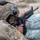TACP Lightning Challenge 2022 A military service member in full combat gear is in a prone or low firing position behind sandbags, aiming a rifle downrange. The soldier is wearing MultiCam/OCP camouflage uniform and a tactical helmet with a reversed American flag patch and communications equipment. The setting appears to be an outdoor training range or field exercise.
