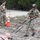 TACP Lightning Challenge 2022 Two military personnel in MultiCam camouflage uniforms are conducting a combat casualty evacuation (CASEVAC) training exercise in an outdoor rocky terrain. One soldier, wearing full tactical gear including a helmet with communications equipment and a plate carrier vest, is dragging a training mannequin using a litter or drag strap. A second soldier stands nearby, apparently observing or timing the exercise.