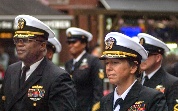 A group of United States Navy personnel in dress uniforms are marching in what appears to be a military parade or ceremonial procession on a city street. Two senior officers are prominently featured in the foreground — a male officer on the left and a female officer on the right — both displaying multiple service ribbons and insignia. Additional Navy enlisted personnel and officers are visible in the background.