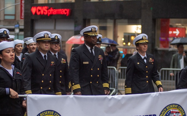 U.S. Navy personnel from the Navy Reserve Center New York City are marching in a parade on a rainy day in an urban setting. Sailors in dress uniforms carry a large white banner identifying their unit. The parade appears to be taking place in New York City, with a CVS Pharmacy storefront visible in the background.