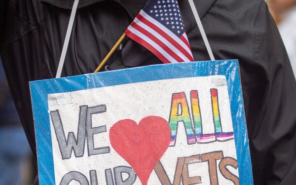 A person wearing a black jacket is displaying a handmade sign that reads 'We Love All Our Vets,' with a small American flag tucked behind it. The word 'ALL' is written in rainbow colors, suggesting inclusivity of all veterans regardless of identity. The setting appears to be an outdoor public gathering or parade.