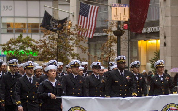 A group of U.S. Navy personnel from the Navy Reserve Center New York City are marching in a parade along a wet city street, suggesting rainy weather conditions. They are dressed in formal Navy dress uniforms with white covers (caps) and dark blue jackets. Two sailors at the front carry a white banner identifying their unit.