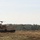 Bull Battery M109A7 Paladin Howitzers take to the field A tan/desert-colored self-propelled howitzer, consistent with an M109 Paladin, is positioned on an open sandy terrain with its long barrel extended horizontally. The vehicle appears to be on a training exercise or operational deployment, situated in front of a dense treeline. The setting and vehicle coloring suggest a military exercise, possibly in a European environment.