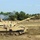 Bull Battery M109A7 Paladin Howitzers take to the field The image shows at least two self-propelled artillery systems in a sandy, open training or exercise area. The foreground vehicle has just fired, as evidenced by visible smoke rising from the rear vehicle behind it and a projectile visible in the upper right of the frame. The setting appears to be a military training range surrounded by pine trees.