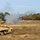 Bull Battery M109A7 Paladin Howitzers take to the field The image shows two self-propelled artillery vehicles, appearing to be M109 Paladin howitzers, firing during what appears to be a military training exercise. A projectile or ejected shell casing is visibly airborne in the upper right portion of the frame, and a large smoke plume is emanating from the lead vehicle's barrel. The setting is an open sandy terrain with tree lines in the background under a partly cloudy sky.