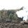 Bull Battery M109A7 Paladin Howitzers take to the field A military exercise or live-fire training event is depicted, showing a tracked self-propelled artillery vehicle firing a round, producing a large white smoke cloud. Several military wheeled vehicles are visible in the background on muddy terrain surrounded by a mixed deciduous and coniferous forest. The scene appears to take place in a temperate European-style environment during autumn.