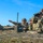 Bull Battery M109A7 Paladin Howitzers take to the field A group of military personnel in camouflage uniforms are conducting what appears to be a field observation or fire direction exercise in an outdoor training environment. One soldier is pointing toward a distant target while others use binoculars to observe, and a green military communications or fire control device is positioned in the foreground. The scene suggests a coordinated observation post or forward observer training exercise.