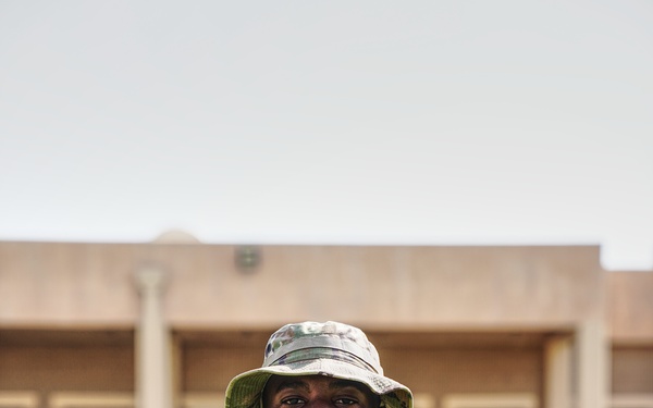 A U.S. Army soldier identified by name tape as 'WATTS' stands at parade rest in an outdoor military installation. He is wearing Operational Camouflage Pattern (OCP) uniform with a boonie hat and displays a Private First Class rank insignia on his chest. The background shows a tan/adobe-style building consistent with a desert military base.