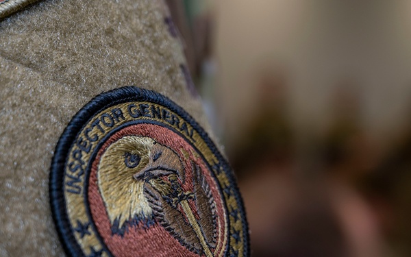 A close-up photograph of a military uniform patch on a camouflage (OCP/MultiCam) uniform. The patch reads 'INSPECTOR GENERAL TEAM' and features an embroidered bald eagle holding what appears to be a sword or lightning bolt, surrounded by stars. The background is blurred, showing indistinct figures in what appears to be an indoor setting.