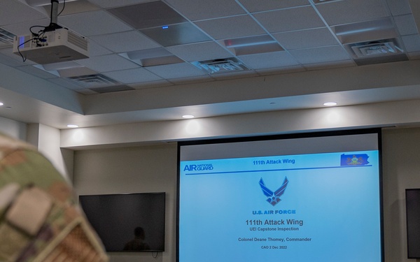 A military briefing is taking place in a conference room at the 111th Attack Wing of the U.S. Air Force Air National Guard. A presentation slide is projected on a large screen showing 'U.S. Air Force 111th Attack Wing UEI Capstone Inspection' with Colonel Deane Thomey listed as Commander, dated December 2, 2022. Military personnel in camouflage uniforms are seated facing the screen while a presenter stands at a podium.