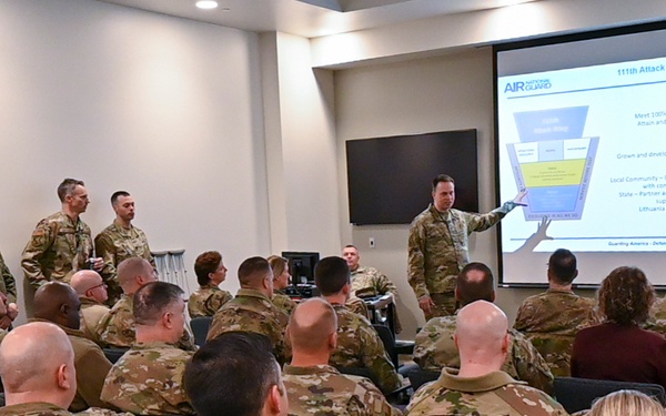 A military briefing is taking place in a conference room where a presenter in camouflage uniform points to a projected slide titled '111th Attack Wing' from the Air National Guard. Approximately 30-40 military personnel in OCP (Operational Camouflage Pattern) uniforms are seated in rows facing the presentation screen. The slide appears to outline organizational goals including 'Operational Excellence,' 'People,' and 'Partnerships.'