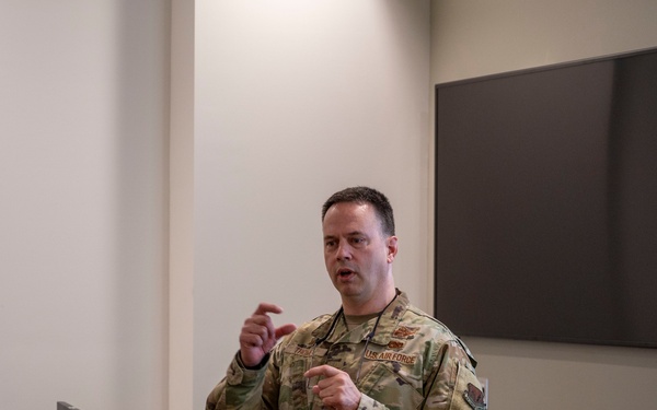 A U.S. Air Force service member in OCP (Operational Camouflage Pattern) uniform is delivering a briefing or presentation in what appears to be a conference or classroom setting. The individual is gesturing with both hands while speaking, with a projection screen visible to the right and a wall-mounted display behind them. A desk with computer equipment is positioned to their left.