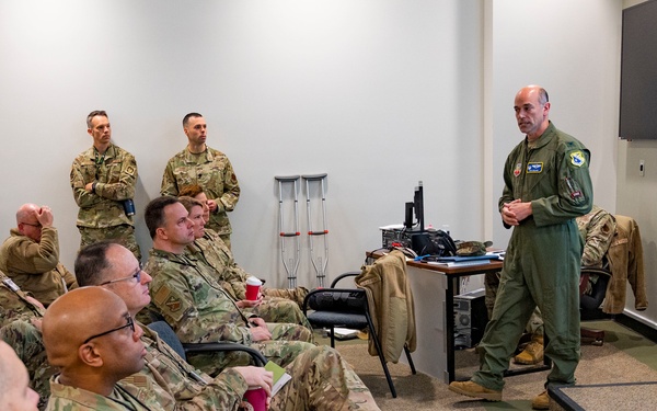 A military briefing or meeting is taking place in a conference room where a presenter in a green flight suit stands before a projected presentation screen. Approximately 15-20 military personnel in OCP (Operational Camouflage Pattern) uniforms are seated and standing as an audience. The setting appears to be a military installation facility, likely a medical or administrative building.