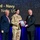 350th Spectrum Warfare Wing Crows awarded at AOC A military award ceremony is taking place on a stage, where a person in U.S. military camouflage uniform is receiving a framed plaque or award. Two civilians in dark suits flank the uniformed recipient, with one presenting the award. The background screen displays text indicating this is a Navy-related award ceremony.