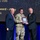 350th Spectrum Warfare Wing Crows awarded at AOC Three individuals are posed on a stage during an awards ceremony. A soldier in U.S. Army camouflage (OCP) uniform is receiving a framed award plaque, flanked by two men in civilian business attire. The background screen displays text referencing an 'Excellence Award – Army' and the name 'Mack, USA'.