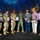 350th Spectrum Warfare Wing Crows awarded at AOC A group of seven individuals pose on a stage during what appears to be a military awards ceremony. Three military personnel in uniform are holding framed plaques or awards, standing alongside four civilians. A large blue digital backdrop with a gold emblem is visible behind the group.
