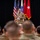 A U.S. Army officer in OCP (Operational Camouflage Pattern) uniform is addressing a large audience of military personnel from a stage or podium area. The speaker stands before multiple flags including the U.S. national flag, a unit or organizational flag, and a red general officer flag with a single white star. The audience, composed of uniformed soldiers, is seated and facing the speaker in what appears to be a formal military briefing or ceremony.