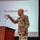 A U.S. Army officer in Operational Camouflage Pattern (OCP) uniform is presenting on a stage at what appears to be a formal military conference or briefing. He is standing near a wooden podium and holding up what appears to be a book or document while speaking. The projection screen behind him identifies the speaker as 'The Adjutant General' with a partially visible name beginning with 'John C. Ha...'