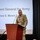 A U.S. Army Brigadier General in OCP (Operational Camouflage Pattern) uniform stands at a wooden podium delivering a presentation. The projection screen behind him identifies the speaker as BG Thomas E. Moore II, serving as Adjutant General for Army. The setting appears to be a formal military briefing or conference.