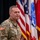 A U.S. Army soldier in OCP (Operational Camouflage Pattern) uniform stands at a podium with a microphone, positioned in front of multiple military and national flags. The soldier's name tape reads 'MOORE' and displays a 10th Mountain Division patch along with a U.S. flag patch on the left sleeve. The setting appears to be a formal military ceremony or official briefing.