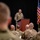 A U.S. Army officer in OCP (Operational Camouflage Pattern) uniform is addressing an audience of fellow soldiers from a wooden podium on a stage. The setting appears to be a formal military briefing or ceremony, with multiple flags displayed behind the speaker. An audience of uniformed soldiers is seated in the foreground, viewed from behind.