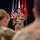 A U.S. Army officer in OCP (Operational Camouflage Pattern) uniform is addressing a military audience while holding a microphone. The speaker stands in front of multiple flags including the American flag and what appear to be general officer flags (red with white stars). The scene appears to be a formal military briefing or ceremony held indoors.