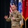 A U.S. Army Colonel is speaking on a stage while holding a handheld microphone, gesturing with his left hand. He is wearing Operational Camouflage Pattern (OCP) uniform with 'U.S. ARMY' tape and a name tape visible on his chest. Several military and U.S. flags are displayed behind him on the stage.