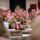 A group of U.S. Army soldiers in MultiCam/OCP camouflage uniforms are seated at a table during what appears to be a military briefing or conference. Three soldiers are prominently visible in the foreground, attentively listening to a presentation. Military unit flags and guidons are visible in the background.