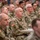 A large group of U.S. Army soldiers in Operational Camouflage Pattern (OCP) uniforms are seated in rows, attending what appears to be a briefing, ceremony, or training event. The soldiers are attentive and facing forward toward an unseen presenter or stage. The setting appears to be an indoor military facility such as a gymnasium or hangar.