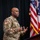 A U.S. Army soldier in MultiCam/OCP camouflage uniform is speaking or presenting near a podium. The soldier wears an Airborne tab and a U.S. flag patch on the left sleeve, and appears to be gesturing with both hands while addressing an audience. Multiple flags, including the U.S. flag and what appears to be military organizational flags, are visible in the background.