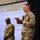 A U.S. Army officer is delivering a presentation at a formal military briefing or leadership event, holding a microphone and gesturing while speaking. A projection screen behind him displays a slide titled 'Message #1: Philosophy' with bullet points referencing Army leadership doctrine (ADP 6-22). A second soldier stands in the background near a podium, with U.S. and organizational flags visible to the right.