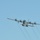 A large four-engine turboprop military transport aircraft, consistent with a C-130 Hercules variant, is photographed in flight against a clear light blue sky. The aircraft is painted white/light gray with distinctive red-tipped wing markings, suggesting a Coast Guard or special mission configuration. Vapor trails or condensation streaks are visible trailing behind the aircraft.