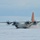 A U.S. Air Force LC-130 Hercules ski-equipped aircraft is operating on a snow/ice runway, likely in Antarctica or the Arctic. The aircraft features distinctive orange tail markings and ski landing gear, with all four turboprop engines running and kicking up snow dust. Green marker flags are visible on the right side, likely delineating the runway boundary.